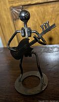 Photo showing a black metal sculpture of a figure playing guitar on a circular base, with a wooden background.