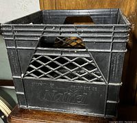 Front-left angle photo of the black plastic milk crate with visible brand name 'J. Nutref' embossed on the side, showing ventilated sides and built-in handles.