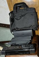 Photo showing a black carrying bag above a black Philips DVD player with remote control on top, placed on a wooden surface against a beige wall.