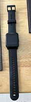 Photo of a black Realme smartwatch on a wooden surface. The watch has a rectangular screen and a black silicone strap with multiple adjustment holes and a buckle.
