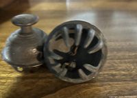 Close-up photo showing the bottom and side of two vintage metal bells on a wooden surface, illustrating their vented bottom design and patina.