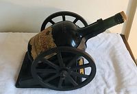 Side view showing the dark glass vintage cognac bottle with an aged label positioned in the black metal cannon-shaped stand with large spoked wheels.