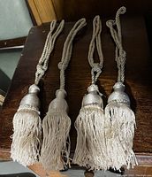 Four beige curtain tassels with twisted cords and fringed ends laid on dark wood surface.