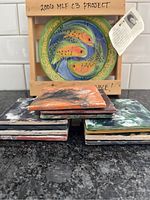 Stack of 7 square ceramic coasters with different abstract patterns, and visible edges showing painted designs and ceramic texture.