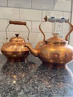 Two copper kettles featuring wooden handles and lids with knobs on a countertop.