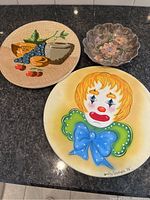 Photo shows three decorative ceramic plates including one with a clown face, one with fruits and vegetables, and a scalloped bowl with floral design.