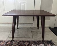 Front view of the vintage mid-century modern small side table showing square top and four tapered legs with brass caps on tile floor.