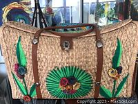 Front view showing large woven straw bag with colorful embroidered floral decorations and leather handles and trim.