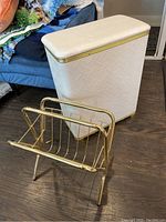 Photo showing white rectangular hamper with gold trim next to brass magazine rack on wooden floor.