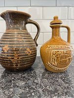 Photo of two vintage pottery pitchers on a counter, one brown with ribbed texture and one mustard yellow with raised designs and inscriptions.