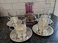 Four glass coffee cups in metal holders with handles, each on a matching metal saucer, alongside a purple glass decanter with round body and cylindrical neck.