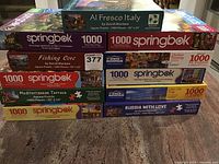 Photo of seven jigsaw puzzle boxes stacked horizontally showing titles and brands clearly.