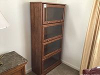 Tall dark wood barrister bookcase with four glass flip doors, brass knobs, standing against a wall near a lamp and curtain.