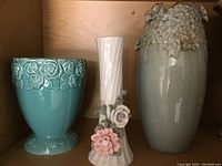 Front view of three vases: teal vase with rose embossed pattern, tall white vase with ceramic flower decorations, tall grey vase with 3D floral appliqués around top