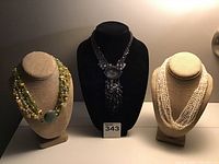 Three costume jewelry necklaces displayed on bust forms, showing the pearl necklace, beaded necklace with green accent bead, and black chain necklace with stone and fringe.