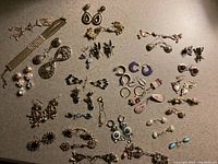 Top-down view of various pierced costume earrings including drop earrings, hoop earrings, filigree designs, rhinestone, bead and enamel embellishments, and a rhinestone mesh bracelet arranged on a flat surface.