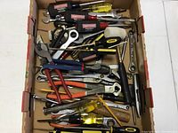 Overview of assorted hand tools in a cardboard box including screwdrivers, wrenches, pliers, cutters, hammer, and scissors.
