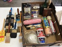 Overview of painting supplies including brushes, rollers, steel wool, masking tape, paint conditioner, and dust mask laid out in a cardboard tray.