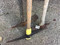 Three picks with different wooden handles placed on concrete ground. One pick has yellow and black grip on handle near metal head.