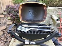Weber Q portable gas grill open showing pig face logo on lid interior and grilling utensils on grate