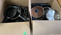 Two large open cardboard boxes filled with assorted pots, pans, and lids showing variety of materials and sizes.