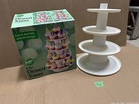 Wilton 4-tier dessert tower fully assembled next to its original packaging box showing the brand and product details, white plastic tiers stacked on a central column.