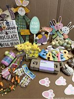 Easter decoration lot with various items arranged on cardboard including wooden bunny and egg hunt signs, plush chicks in packaging, pastel decorative eggs, colorful wooden letters spelling EASTER, carrot wreath, paper bunny footprints, and small packaged stickers.