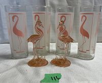 Lot showing four vintage flamingo drinking glasses and two pink glass flamingo figurines arranged together on cloth background.
