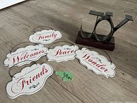 Six decorative white metal Christmas signs laid out on floor, each with a different holiday saying and black wire hangers attached. Signs read 'Friends,' 'Welcome to Our Home,' 'Family,' 'Peace is the Heart of Christmas,' 'Wonder of Christmas Children' plus a metal holder stand accessory.