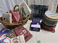 Photo showing woven basket with multiple unopened packs of decorative paper napkins, holiday crackers, and a box of plastic cutlery alongside a stack of colorful striped paper plates.