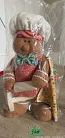 Front and side view of the plush gingerbread man baker sealed in original plastic wrapping, showing its detailed features and accessories.