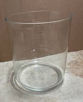 Front view of a large clear glass vase placed on a floor with a brown background, shows full height and shape.