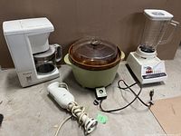 Full view of vintage small kitchen appliances group: crockpot, coffee maker, hand blender on the floor, and full-size blender with glass container on the right.