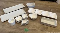 All white ceramic servingware items grouped on wooden floor including various trays, dishes, bowls, and spoons