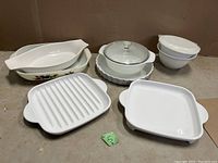 Full view of the white ceramic CorningWare cookware set on surface showing different shapes including grill pan, oval and rectangular dishes, round casserole with glass lid, and nested bowls with lids.