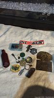 Photo showing a collection of vintage items including badge buckles, tokens, a leather pouch, miniature spoon, figurine, and other small collectibles spread on a white cloth.