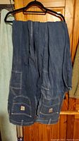 Two pairs of blue denim Carhartt jeans hanging on a hanger, showing reinforced pockets and Carhartt logo patch.