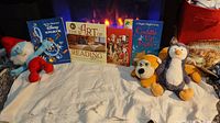 Photo showing all four children's books and two Kellytoy plush toys (Grandpa Smurf and penguin) laid out on a white cloth in front of a fireplace.