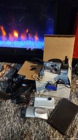 Photo shows vintage Olympus camera in original box with strap and cords, black calculator, and silver digital camera with black case on carpeted floor.