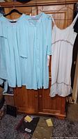 Three garments hanging: two light blue items (nightgown and house coat) and one white nightgown with floral embroidery and ruffled bottom