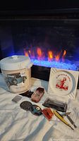 Full lot shown with fireplace background. Items include vintage white tin container with VW van graphic, two Zippo lighters, amber cologne bottle, white ashtray, two belt buckles, straight razor, screwdriver tool, and keychain objects.