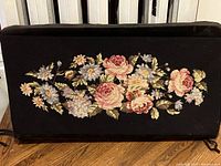 Full view of black velvet piano bench cover with large floral needlepoint embroidery in the center, placed on wooden floor.