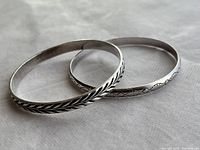 Two silver bracelets with different engraved designs shown together on a white fabric background.