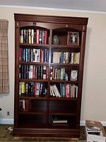 Front view of one cherry wood bookshelf filled with books and some decorative items, showing glass inlay on upper shelves and crown molding.