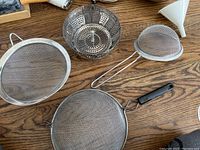 Various metal mesh sifters and strainers, expandable steamer basket, plastic funnel, black handled utensil on wood table