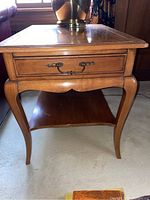 Wood end table showing drawer with metal handle and curved legs, surface shows some wear.