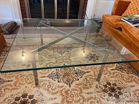 Square glass coffee table with silver metal legs viewed from above showing clear glass top and silver X-shaped support frame.