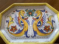 Large octagonal ceramic serving bowl with yellow interior and two stylized sea monsters facing each other, surrounded by floral and ornamental designs in blue, yellow, red, and green colors.
