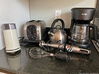 Full view of all six kitchen appliances on black granite counter including Mr. Coffee coffee maker, Braun coffee grinder, stainless steel toaster, stainless steel tea kettle, stainless stove-top coffee maker, and Cuisinart immersion blender with whisk attachment.