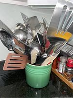 Photo showing a green ceramic crock containing various large kitchen utensils including spatulas, ladles, slotted spoons, and forks.